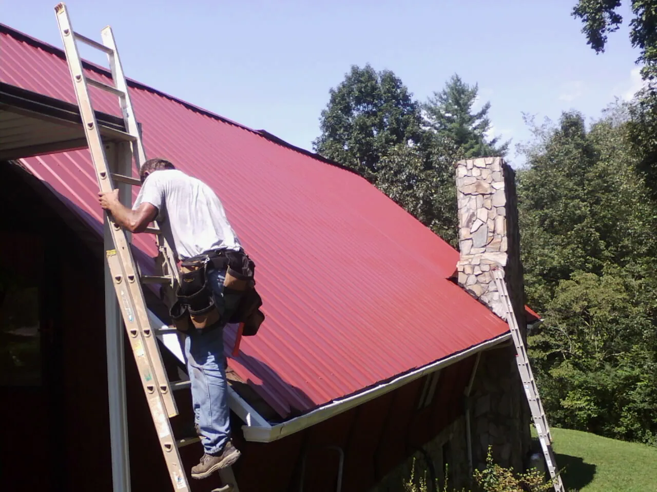 Roofer on a ladder beside a red metal roof and stone chimney — Soffit & Fascia Repair by Roofing Brothers in New Kensington